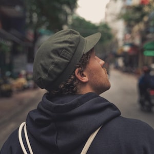 Op de afbeelding: Een groene pet met een korte klep. De pet wordt gedragen door een persoon die een marineblauwe hoodie draagt. De persoon kijkt omhoog, met een vage straatscène op de achtergrond.
