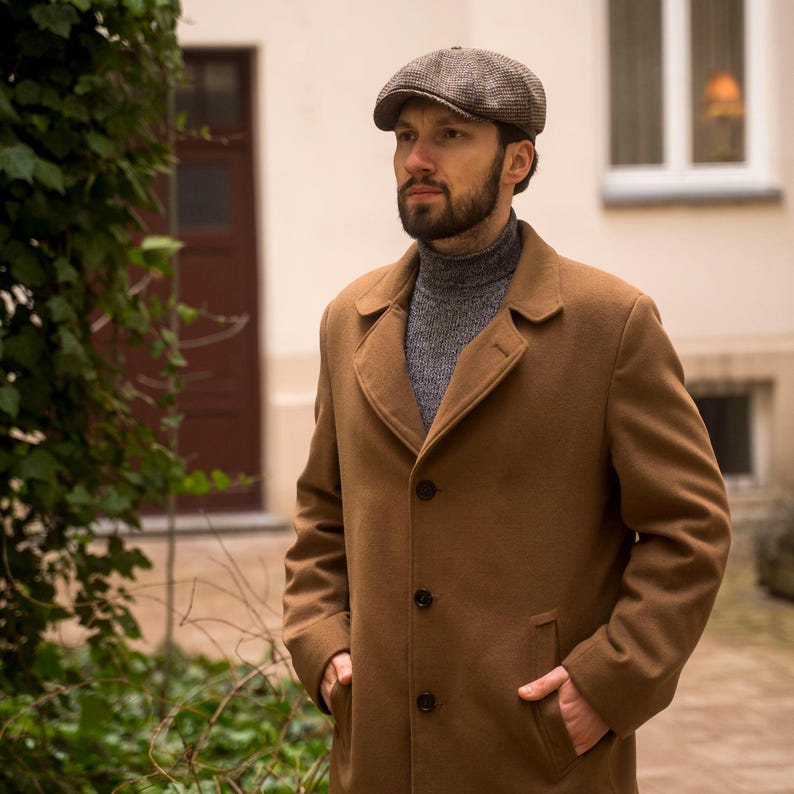 Puede incluir: Un hombre con un abrigo de lana marr&oacute;n y un jersey de cuello alto gris debajo. Lleva una gorra de tweed marr&oacute;n y gris.