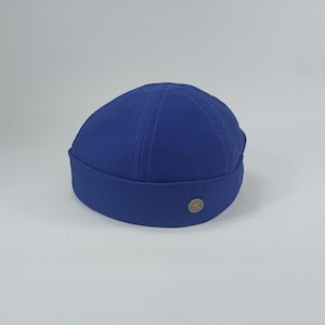 May include: A cobalt blue skull cap with a rounded crown and a folded brim. The cap has a small, circular metal emblem on the side. The hat is made of a durable fabric and is suitable for various casual outfits.
