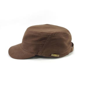 May include: A brown military-style cap with a short brim. The cap is made of a solid, textured fabric and features a small, rectangular metal emblem on the side. The cap has a structured crown and a slightly curved brim.