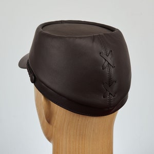 May include: A brown leather cap with a stitched X pattern on the back. The cap has a short brim and a button on the side.