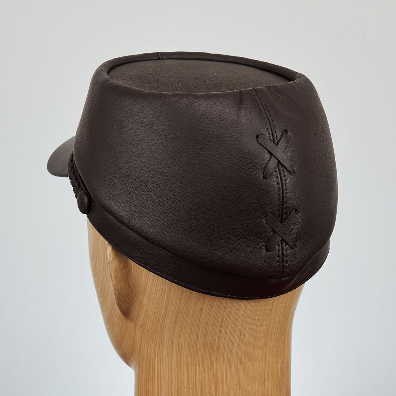May include: A brown leather cap with a stitched X pattern on the back. The cap has a short brim and a button on the side.