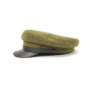 Op de afbeelding: Olijfgroene pet met een donkerbruin leren klep en band. De pet heeft een afgeronde kroon en een kleine, zwarte knoop aan de zijkant. De hoed is gemaakt van een textuurstof en staat tegen een witte achtergrond.