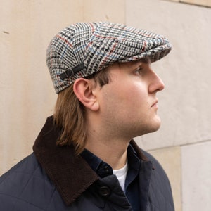 May include: A patterned newsboy cap in a classic style. The cap features a black, white, red, and blue plaid design. It has a small, black leather strap detail on the side. The cap is made of a textured fabric.