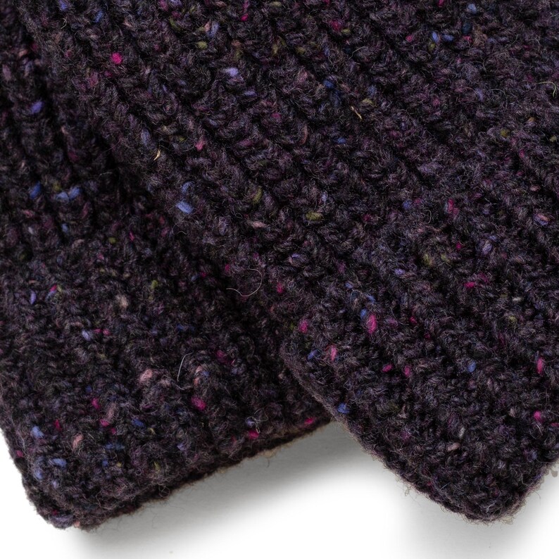 May include: Close-up of a dark purple knitted fabric with a ribbed texture. The fabric has flecks of blue, pink, and green throughout, adding visual interest. The material appears to be soft and warm, suitable for winter wear.