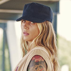 May include: A navy blue military-style cap with a short brim. The cap is worn by a person with long blonde hair and a tattoo on their arm. The person is wearing a beige sweater.