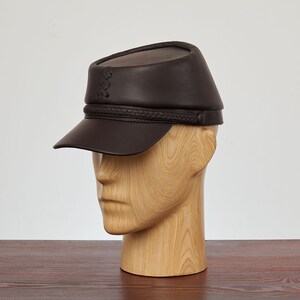 May include: A brown leather cap with a braided band and a small stitched design on the top. The cap has a short brim and is worn on a wooden mannequin head.