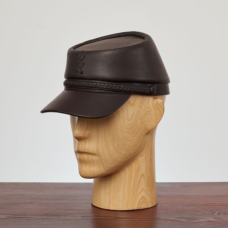 May include: A brown leather cap with a braided band and a small stitched design on the top. The cap has a short brim and is worn on a wooden mannequin head.