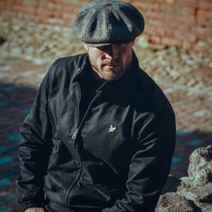 Puede incluir: Una chaqueta negra con cierre de cremallera y un peque&ntilde;o logotipo bordado en el pecho. La chaqueta la lleva puesta una persona que lleva una gorra de tweed de estilo newsboy.