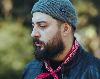 May include: A gray beanie with a small blue tag is worn by a person with a full beard. A red paisley bandana is tied around the neck, and a black leather jacket is visible. The background is blurred with green and brown tones.