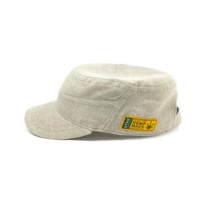 May include: A light beige military-style cap made of hemp fabric. The cap features a short brim and a flat top with a structured design. A yellow and green patch on the side reads "HEMP MADE".