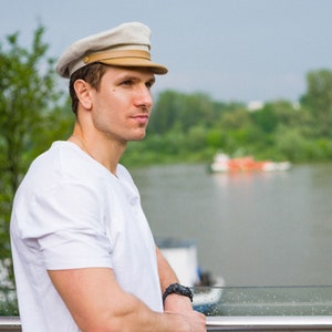 May include: A man wearing a white t-shirt and a light beige cap with a brown brim. The cap has a button on the top and a brown band around the brim. The man is standing in front of a body of water.