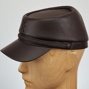 May include: A brown leather cap with a braided band and a button on the side. The cap has a short brim and a rounded crown.