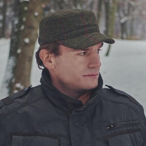 May include: A green tweed cap with a flat top and a short brim. The cap has a subtle red plaid pattern and is worn by a person. The person is wearing a black jacket. The background is a snowy outdoor scene.