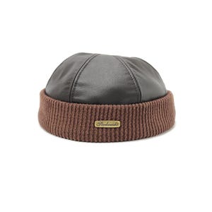 Puede incluir: Un gorro marrón y negro con la parte superior de cuero y un borde de punto acanalado. El gorro presenta una pequeña etiqueta rectangular dorada con el nombre "Sterkowski" en la parte delantera.