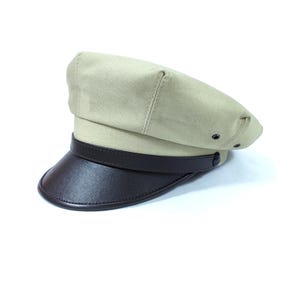 Op de afbeelding: Een beige pet met een donkerbruin, leerachtig vizier. De pet heeft een gestructureerd ontwerp met een band rond de kroon. De klep is gebogen en de pet heeft een klassieke, vintage stijl.