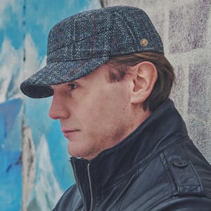May include: A gray tweed cap with a short brim and a small gold emblem on the side. The cap has a plaid pattern with red and blue accents. The person in the image is wearing a black leather jacket.