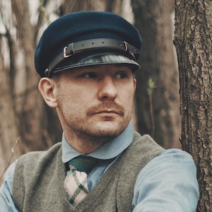 Op de afbeelding: Een marineblauwe pet met een leren klep en riem. De pet wordt gedragen met een lichtblauw overhemd met kraag, een grijze trui en een stropdas met patroon. De afbeelding is buiten gemaakt met een vage achtergrond.