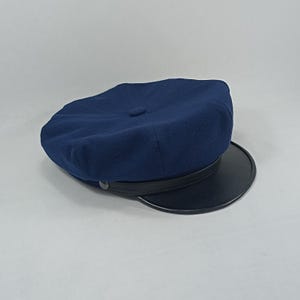 May include: A navy blue wool cap with a black leather brim. The cap has a button on the top and a stitched seam around the brim.