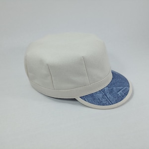 May include: A white canvas cap with a blue denim brim. The brim has a white trim.