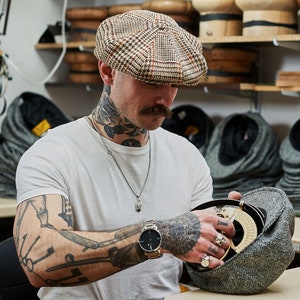 K&ouml;nnte beinhalten: Ein Mann mit T&auml;towierungen tr&auml;gt eine braune und schwarze gemusterte Newsboy-Kappe und eine silberne Kettenkette. Er h&auml;lt eine graue und schwarze Tweed-Newsboy-Kappe in seinen H&auml;nden. Er tr&auml;gt ein wei&szlig;es T-Shirt und eine schwarze Uhr.