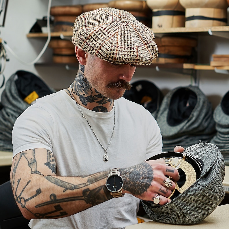 K&ouml;nnte beinhalten: Ein Mann mit T&auml;towierungen tr&auml;gt eine braune und schwarze gemusterte Newsboy-Kappe und eine silberne Kettenkette. Er h&auml;lt eine graue und schwarze Tweed-Newsboy-Kappe in seinen H&auml;nden. Er tr&auml;gt ein wei&szlig;es T-Shirt und eine schwarze Uhr.