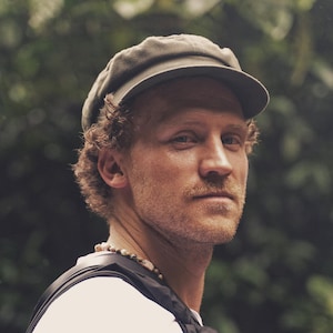 Op de afbeelding: Een man met een groene pet en een wit shirt. Hij heeft krullend bruin haar en een snor. Een kralenketting hangt om zijn nek. De achtergrond is een vage groene bladermassa.