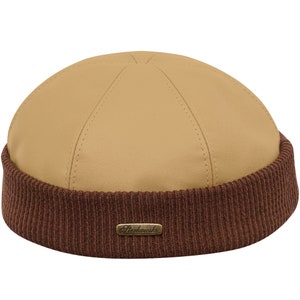 Op de afbeelding: Een beige leren beanie met een bruin gebreid band. De beanie heeft een klein, goudkleurig label met de tekst "Sterkowski" erop.