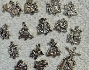 May include: A set of 15 silver-toned metal charms depicting scenes from the Stations of the Cross. Each charm features a detailed depiction of a different station, including Jesus carrying the cross, Jesus falling, and Jesus being nailed to the cross.
