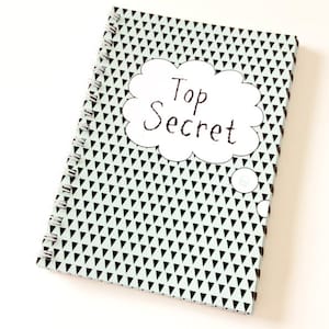 Könnte beinhalten: Ein spiralgebundenes Notizbuch mit einem Cover, das ein Muster aus schwarzen Dreiecken auf hellgrünem Hintergrund zeigt. Ein weißes Wolken-Grafik mit dem Text "Top Secret" ist auf dem Cover zentriert.