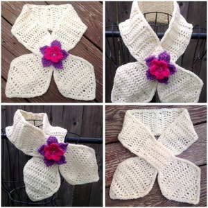 May include: A white crocheted infinity scarf with a purple and pink flower accent. The scarf is folded in a way that creates a bow shape.