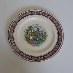May include: A white ceramic plate with a gold rim and a pink and green floral border. The center of the plate features a colorful illustration of a cottage with the text "The Old Mill".