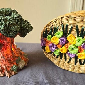 May include: A decorative tree with a green leafy canopy and a brown, textured trunk. The tree is illuminated from within, casting a warm glow. Next to the tree is a woven basket with a floral design made of colorful ribbons.