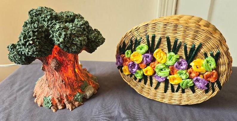 May include: A decorative tree with a green leafy canopy and a brown, textured trunk. The tree is illuminated from within, casting a warm glow. Next to the tree is a woven basket with a floral design made of colorful ribbons.