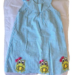 May include: A sleeveless, light blue and white gingham dress with frog appliqués. The dress features pink bows on the shoulders and at the bottom, with three frog designs. The frogs are yellow with black eyes and a pink bow.
