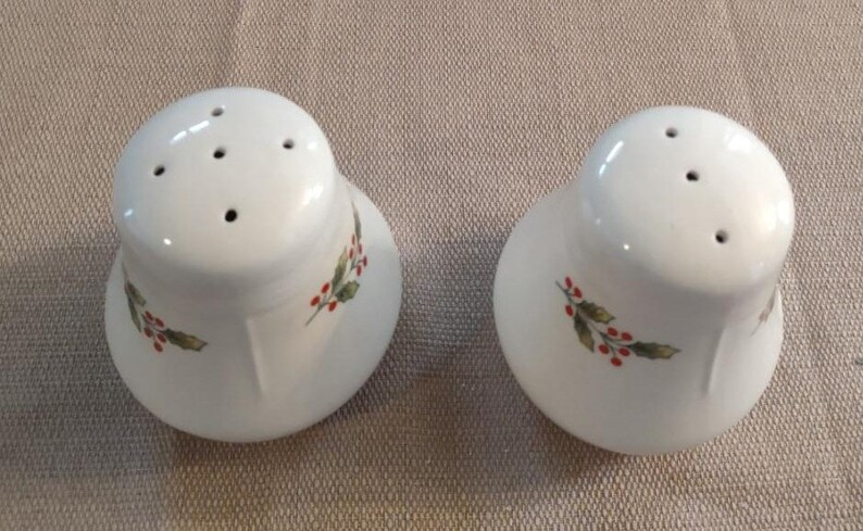 May include: Two white ceramic salt and pepper shakers with a holly and berry design. The shakers have four holes on the top for dispensing salt and pepper.