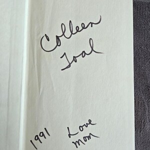 May include: Open book page with handwritten inscription. The text reads "Colleen Joal" above and "1991 Love mom" below, all in black ink. The page is off-white, with a red edge.