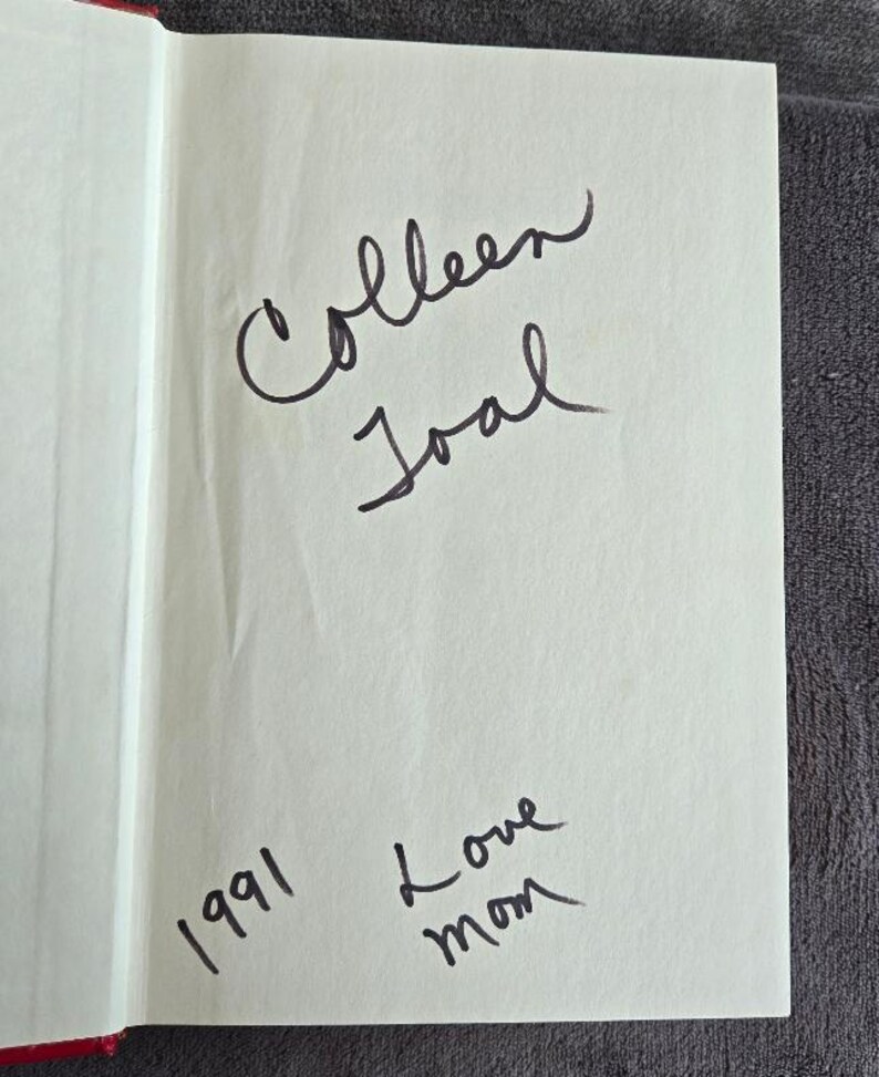 May include: Open book page with handwritten inscription. The text reads "Colleen Joal" above and "1991 Love mom" below, all in black ink. The page is off-white, with a red edge.
