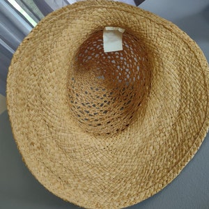 Puede incluir: Un sombrero de paja de ala ancha con un dise&ntilde;o tejido. El sombrero es de color marr&oacute;n claro y tiene una etiqueta blanca adherida al interior.