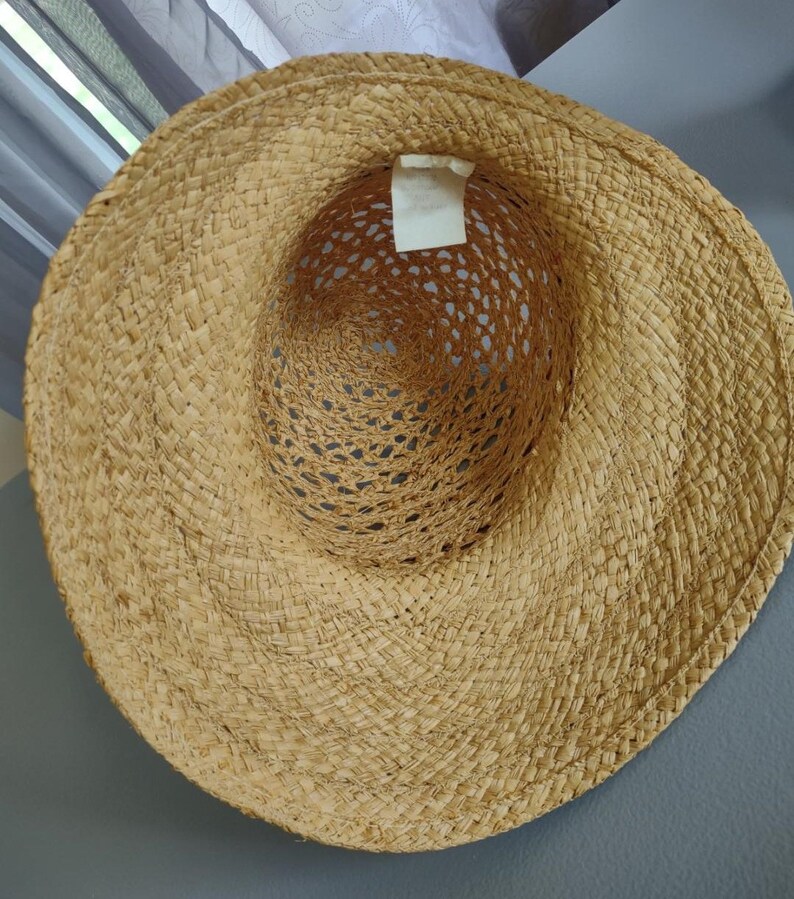 Puede incluir: Un sombrero de paja de ala ancha con un dise&ntilde;o tejido. El sombrero es de color marr&oacute;n claro y tiene una etiqueta blanca adherida al interior.