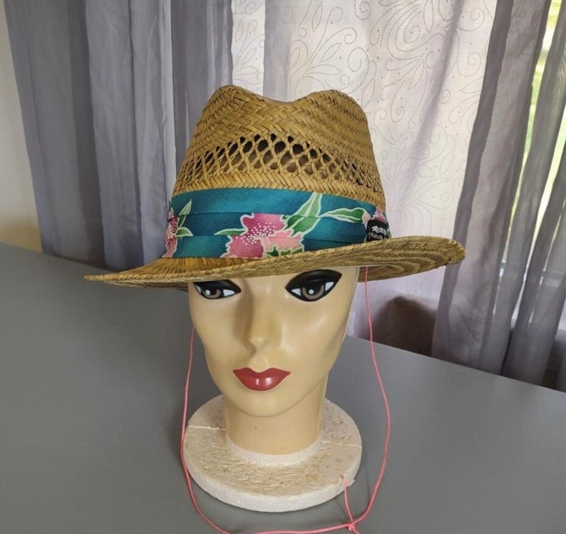 Peut inclure: Un chapeau de paille avec un ruban floral turquoise et rose. Le chapeau a un design tiss&eacute; et une ficelle rose attach&eacute;e.