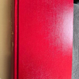 May include: A hardcover book with a bright red cover. The spine is visible, with gold lettering. The pages are a light cream color. The book is resting on a gray surface.