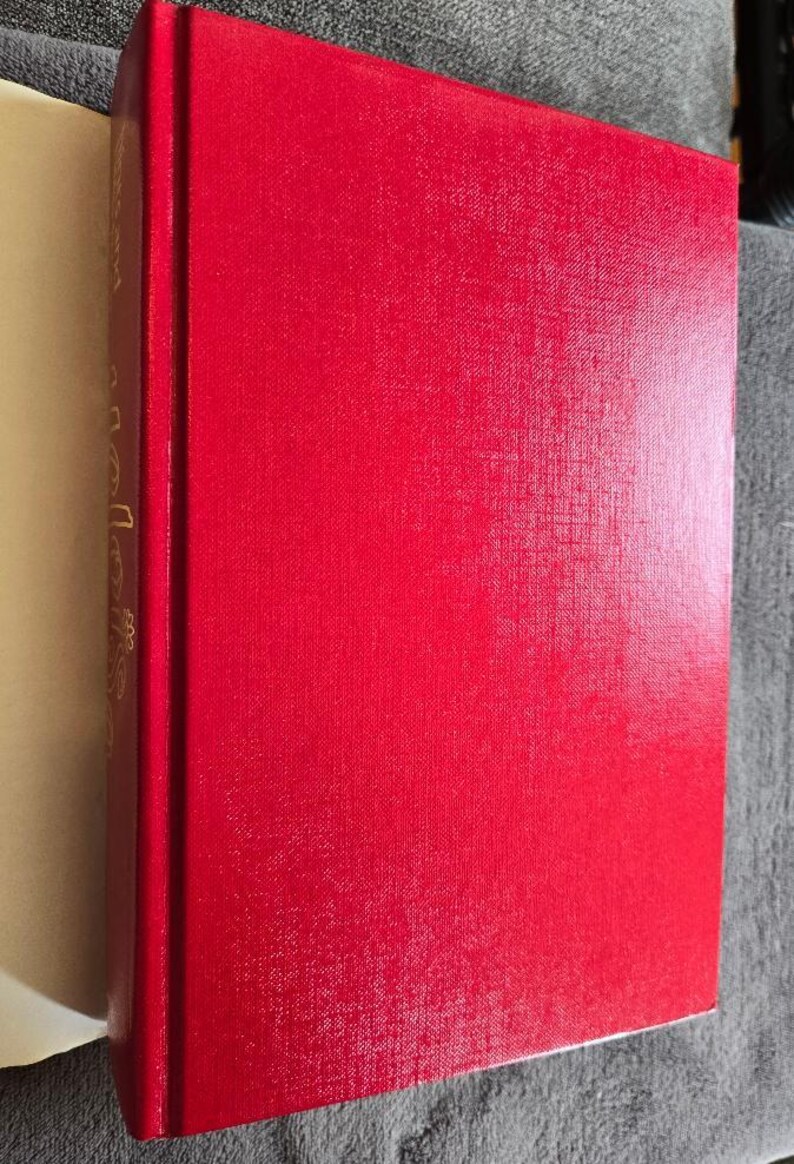 May include: A hardcover book with a bright red cover. The spine is visible, with gold lettering. The pages are a light cream color. The book is resting on a gray surface.