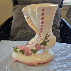 May include: A white ceramic boot-shaped vase with pink floral designs and pink buttons. The vase is decorated with pink roses and green leaves.