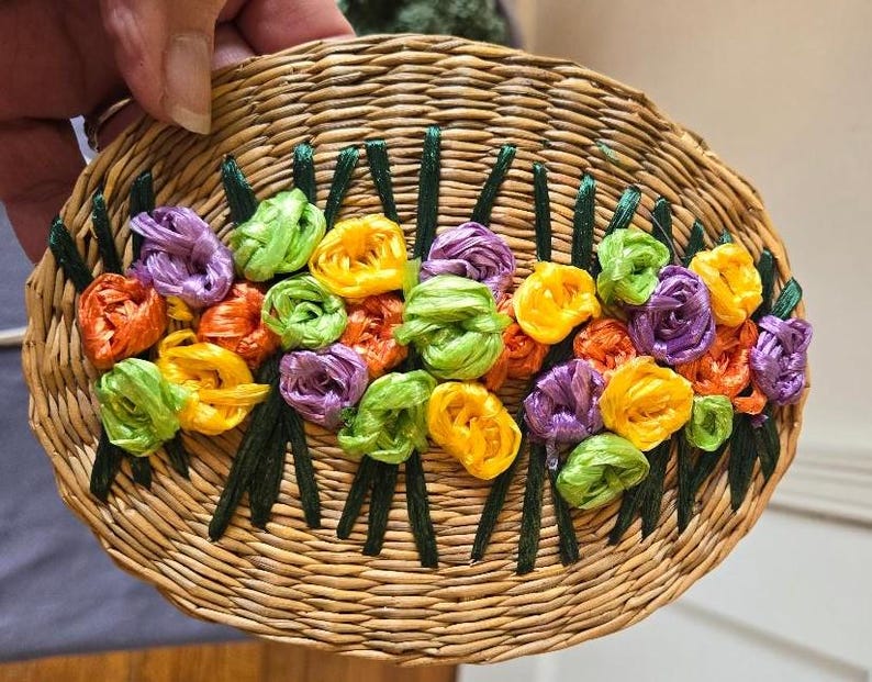 May include: A woven oval-shaped coaster with a design of colorful fabric flowers in shades of yellow, green, purple, and orange.