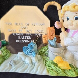 May include: A figurine of a child holding a fishing rod with a blue fish on the line. The child is standing next to a bucket with a blue fish inside. The figurine is on a base with a green grass and flower design. A silver plaque with the text "FOR BLUE OF STREAM AND BLUE OF SKY LORD. THANK YOU FOR EASTER BLESSINGS." is behind the figurine.