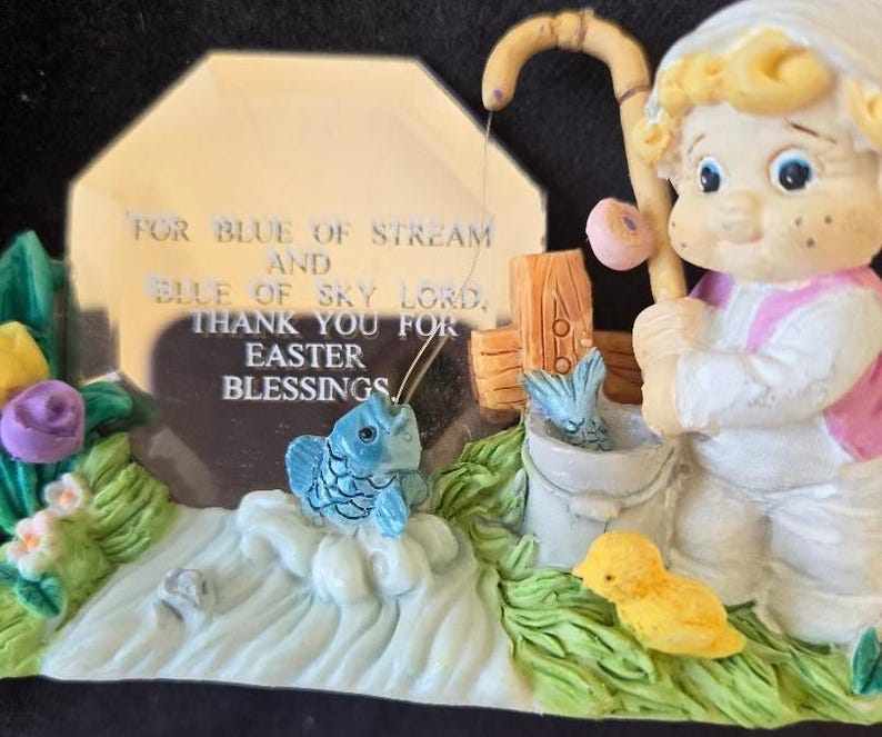 May include: A figurine of a child holding a fishing rod with a blue fish on the line. The child is standing next to a bucket with a blue fish inside. The figurine is on a base with a green grass and flower design. A silver plaque with the text "FOR BLUE OF STREAM AND BLUE OF SKY LORD. THANK YOU FOR EASTER BLESSINGS." is behind the figurine.