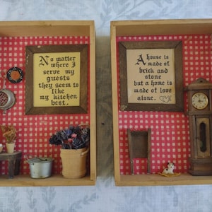 May include: Two wooden shadow boxes with red and white checkered backgrounds. The left box features a framed quote about kitchens and a miniature kitchen scene with a pot, a stool, and a strainer. The right box features a framed quote about home and love, a miniature grandfather clock, a chair, and a small dog figurine.