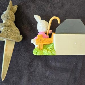 May include: Two ceramic bunny figurines. One bunny is sitting on a bench with a cane and the other bunny is a cake topper with a silver pick.