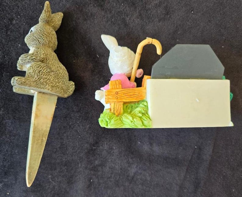 May include: Two ceramic bunny figurines. One bunny is sitting on a bench with a cane and the other bunny is a cake topper with a silver pick.